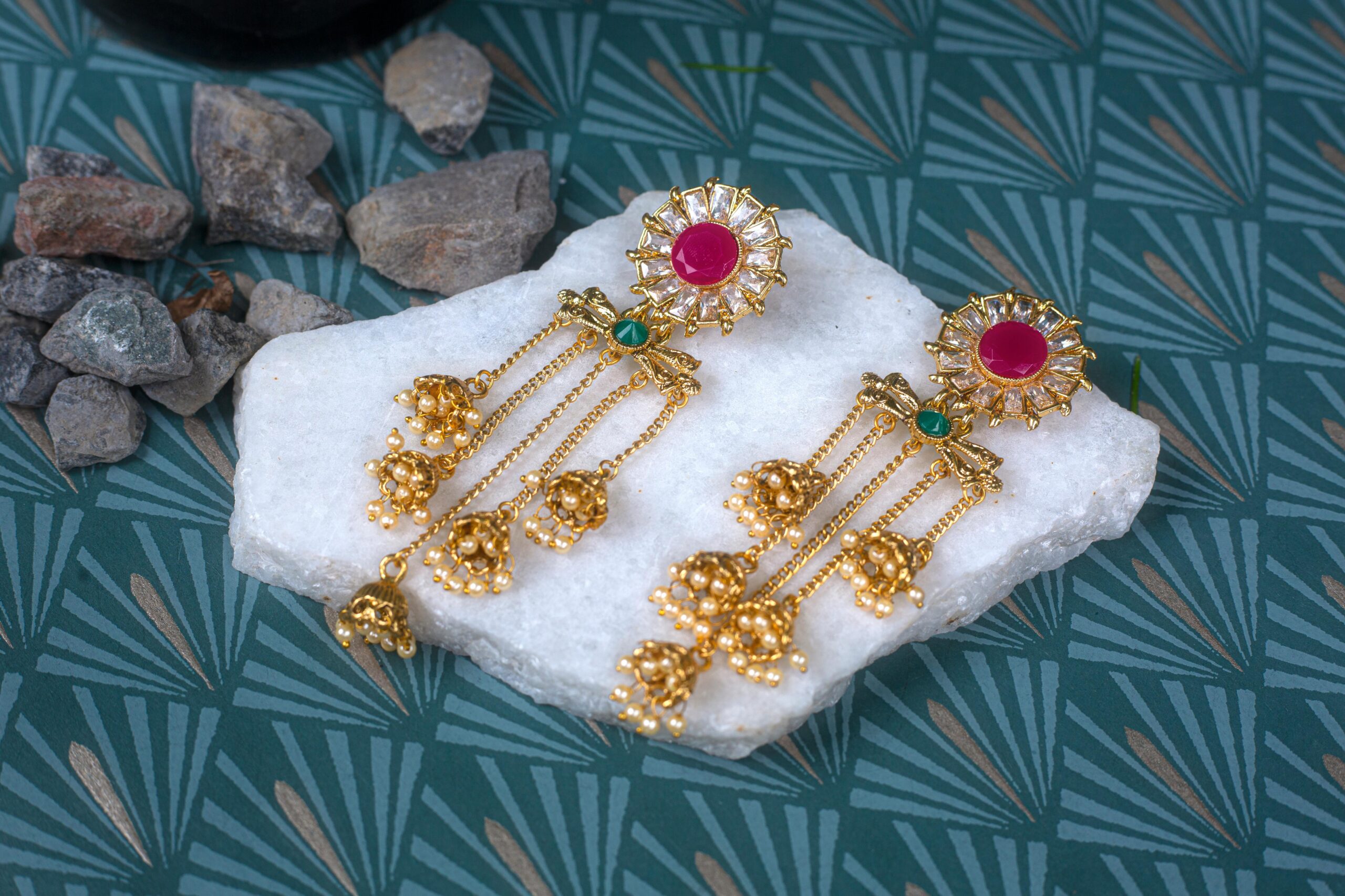 Luxurious gold earrings with ruby and emerald stones displayed on white marble, exuding elegance and tradition.